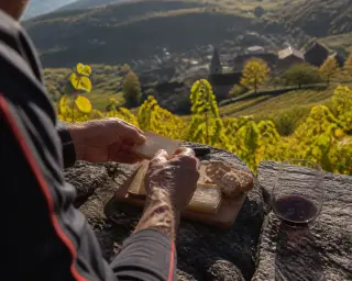 Hiker's hands preparing a simple picnic with cheese, bread, and wine on a stone wall overlooking autumn vineyards and a village in soft afternoon light
