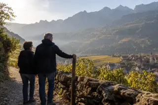 Couple pausing by a vineyard path above a Savoie village at dawn, looking toward mountains in soft, golden light with mist and patchwork slopes visible.